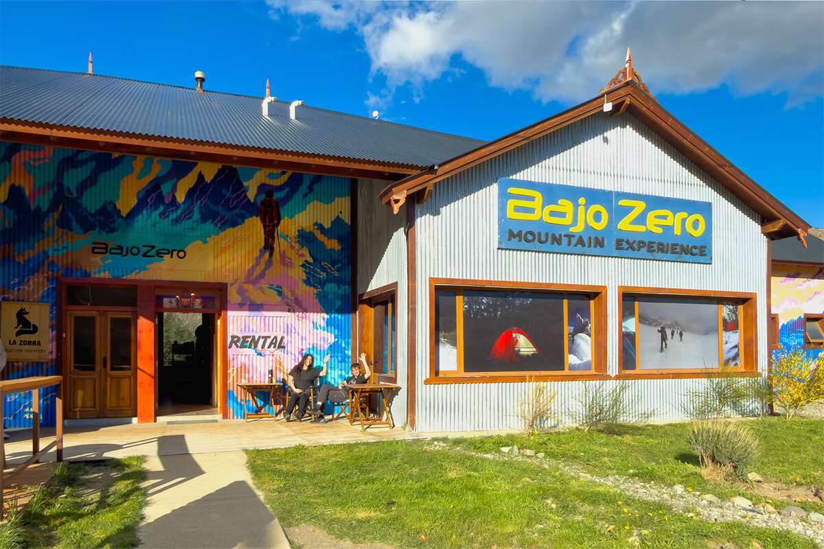 Fachada de Bajo Zero con un mural de montañas, el cartel de alquiler y personas sentadas en la vereda