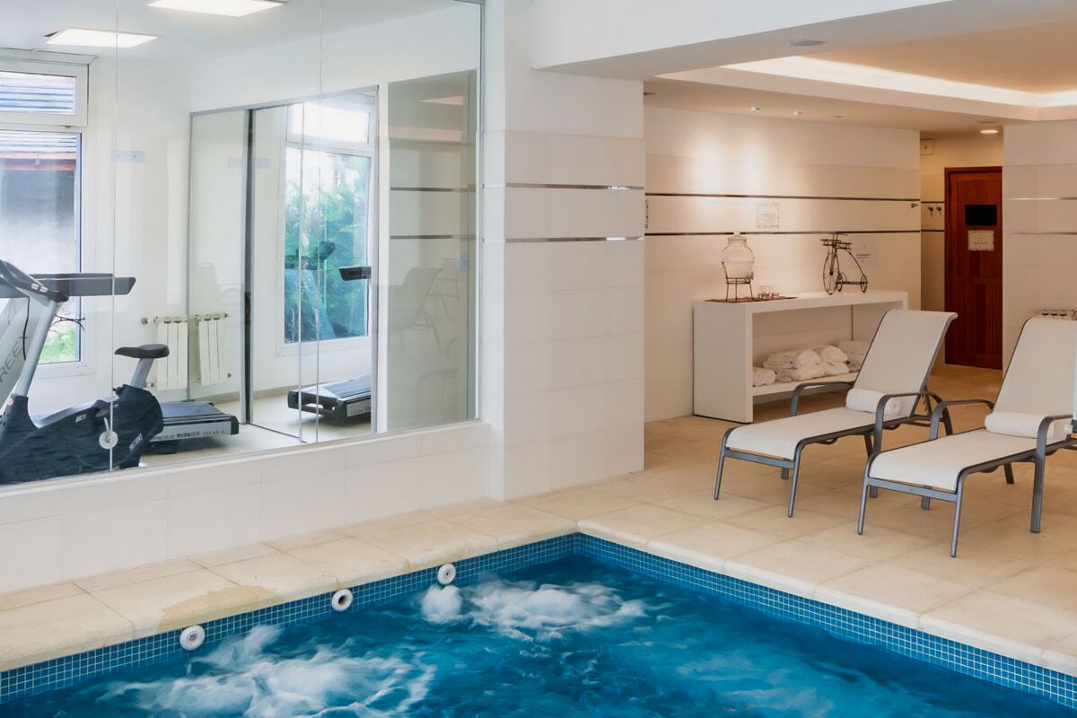 Indoor pool with hydromassage, white loungers and the gym behind a glass wall