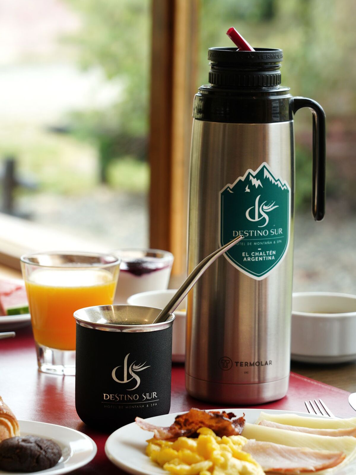 Thermos and mate with the Destino Sur logo next to scrambled eggs, cold cuts and orange juice
