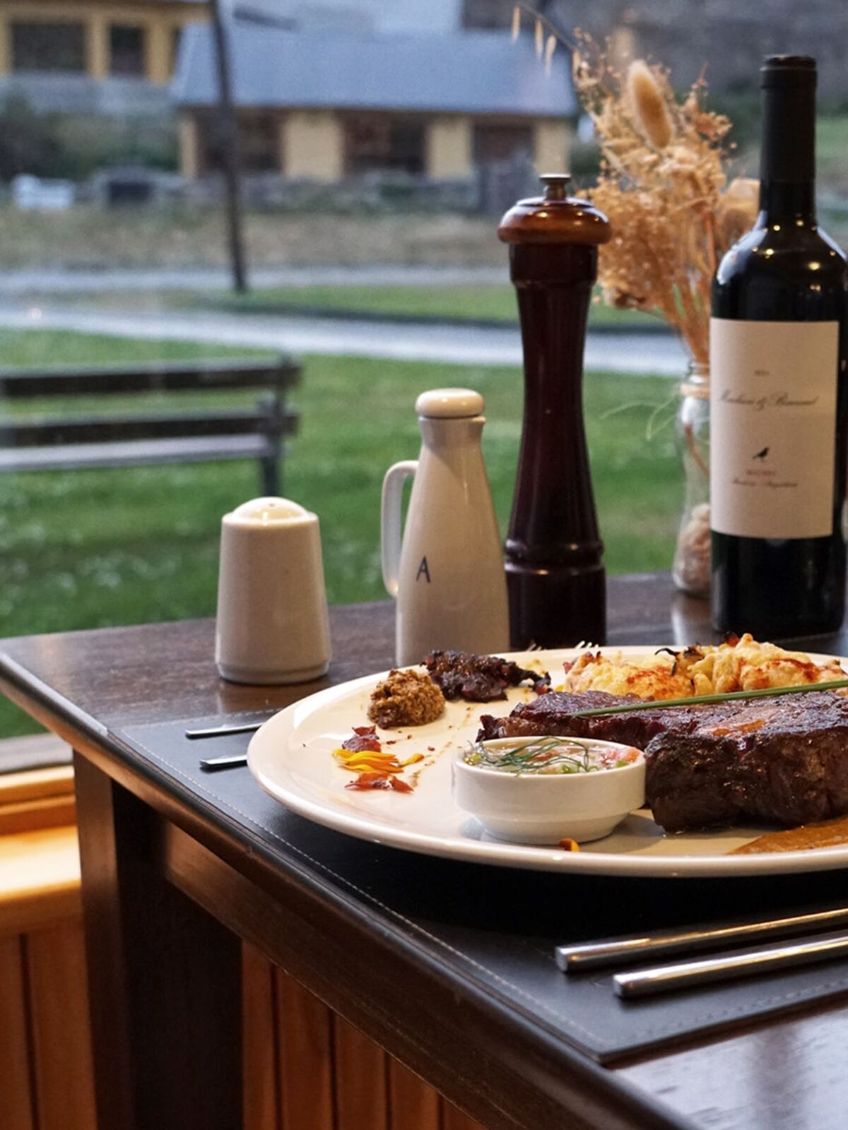 Plate of steak and a bottle of red wine by the window, with the garden and village houses behind