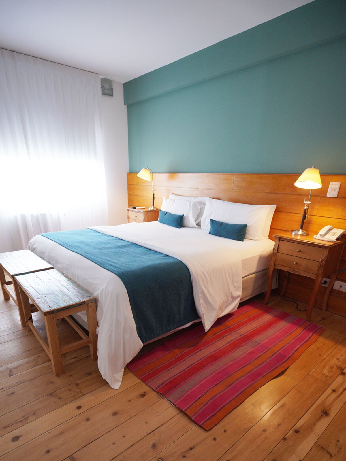 Room with a turquoise wall, a double bed with a blue throw, a striped rug and wooden floors