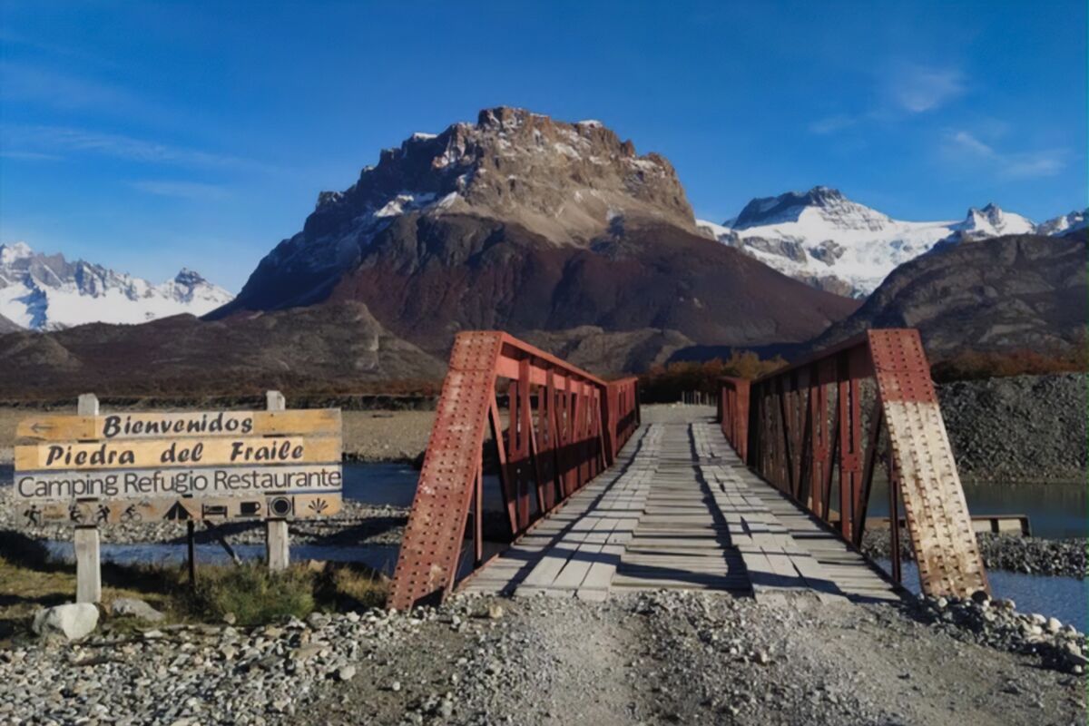 Puente de hierro rojo y cartel de bienvenida a Piedra del Fraile, con montañas detrás