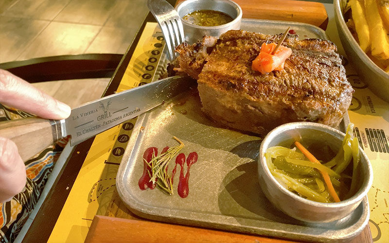 Bife grueso servido en tabla con papas, salsas y un cuchillo de mesa