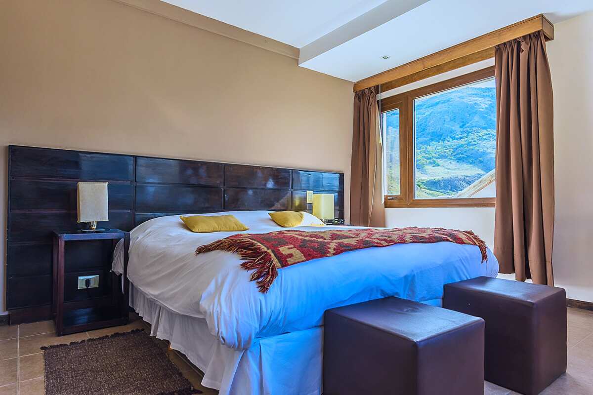 Room with a double bed, dark wooden headboard, two ottomans and a window with mountain views