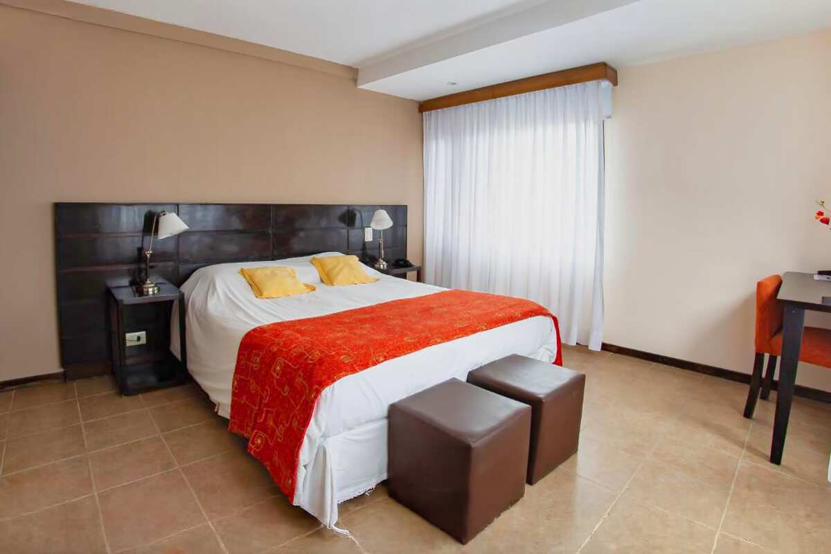 Room with a double bed, two ottomans, a desk and white curtains