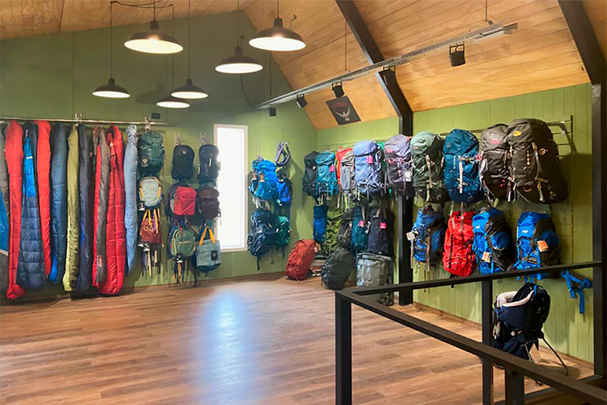 Planta alta con bolsas de dormir y mochilas grandes colgadas bajo un cartel de Osprey, y una mochila porta bebé
