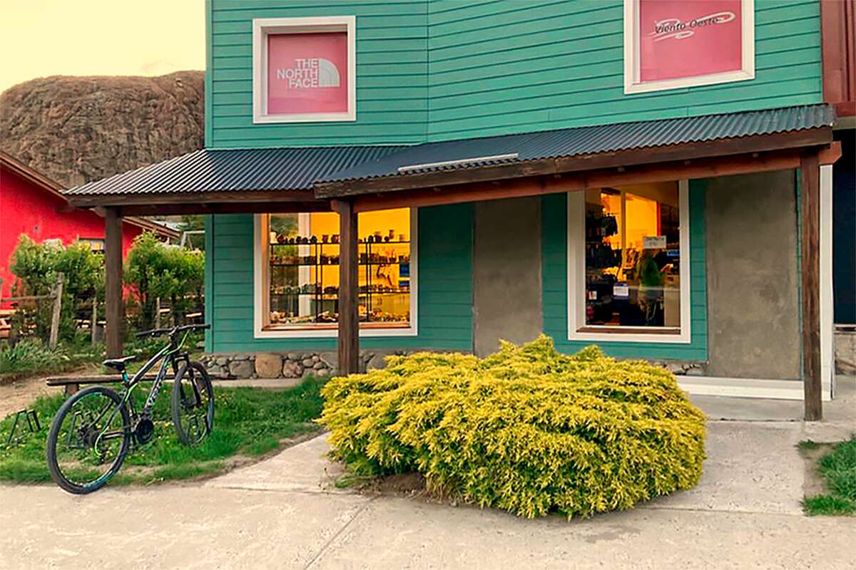 Frente de madera verde con galería, una bicicleta apoyada y carteles de The North Face y Viento Oeste