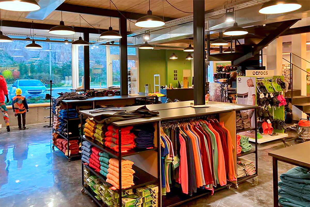 Salón con ventanales, mesas de ropa doblada y exhibidor de calzado Crocs